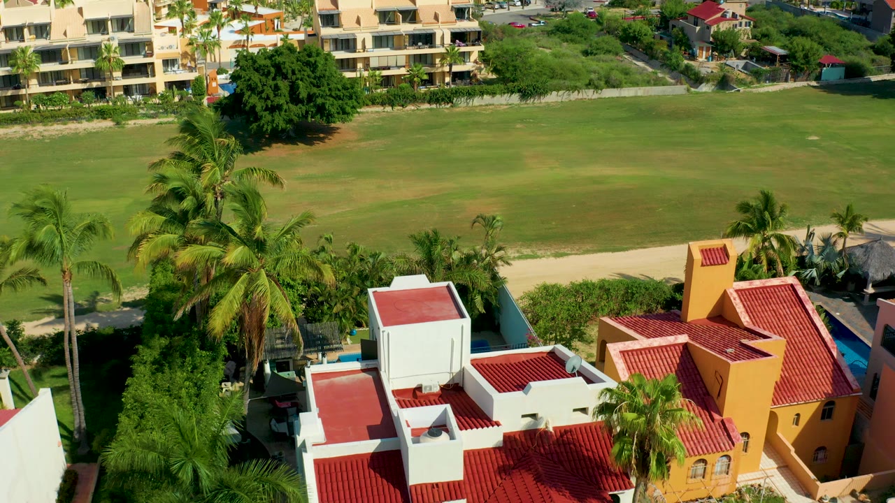 Aerial frame in San Jose del Cabo showing red roofed white and yellow villas below a broad green lawn, with palms in the foreground and no visible people.
