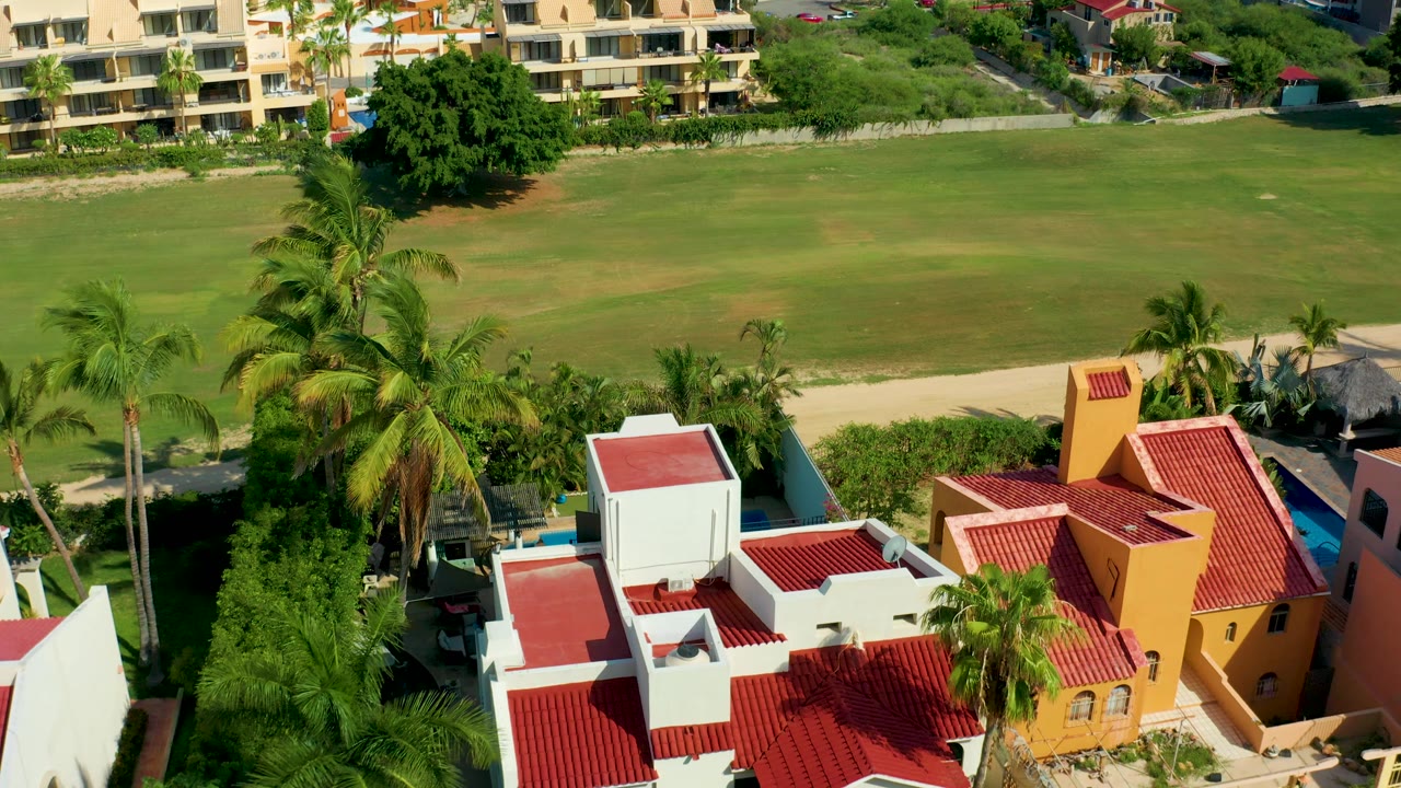San Jose del Cabo aerial frame with a yellow villa, white roof sections, palms, and the large grassy field stretching across the background, with no visible people.