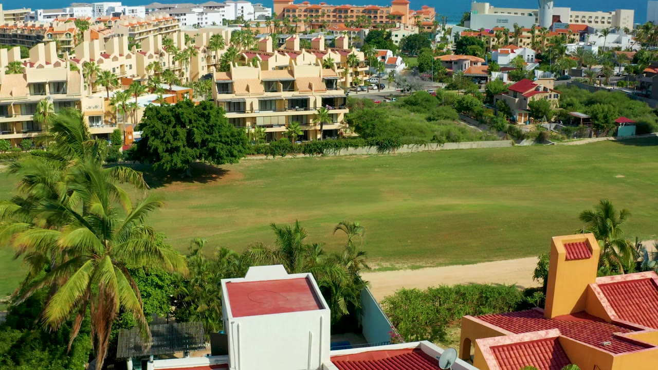 Opening frame of San Jose Del Cabo, Mexico, showing red-tiled rooftops and palms in front of a green field and cream apartment buildings, with no visible people.
