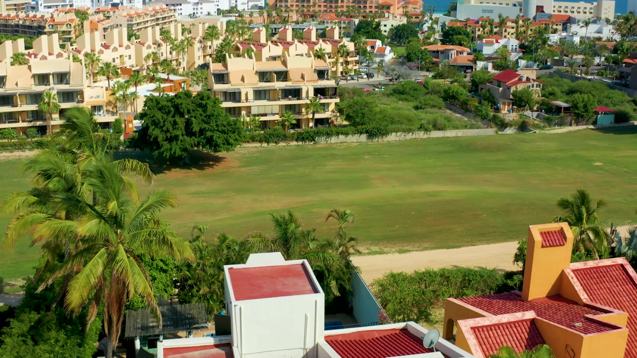 San Jose Del Cabo residential frame with palms and red rooflines below, an uninterrupted green field across the center, and cream apartment blocks in the background, with no visible people.