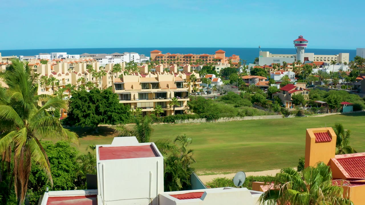 Early frame in San Jose del Cabo showing palms beside white and terracotta foreground roofs, a large lawn, dense residential buildings, distant ocean, clear sky, and no visible people.