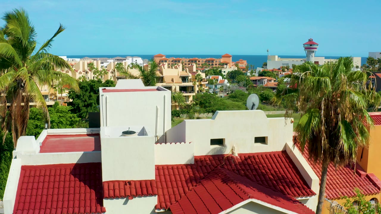 White rooftop structure, red roof tiles, palms, town buildings, and distant ocean.