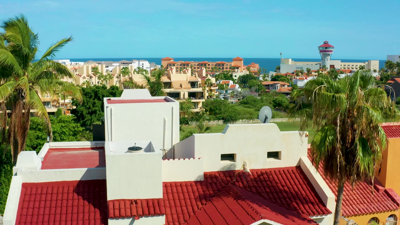 San Jose del Cabo rooftop angle showing red roof tiles and white walls in the foreground, palms partly covering the right side, residential blocks and ocean beyond, and no visible people.