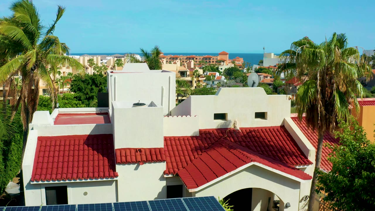 Opening frame of a San Jose del Cabo white stucco building with red tile roofs, palms on both sides, a strip of solar panels at the bottom, distant buildings, ocean, and no visible people.