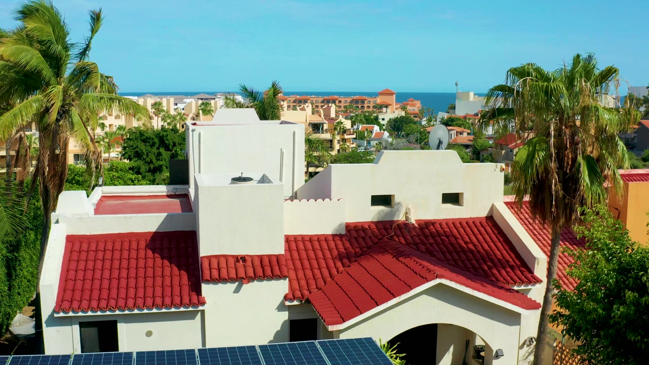 At 0:02, the San Jose del Cabo building appears slightly lower in frame, showing more roof surface, tall palms, distant tan and white buildings, the blue ocean, and no visible people.