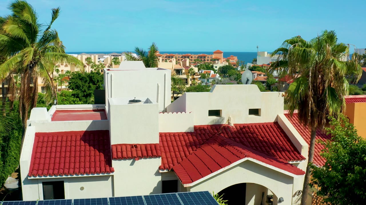 Solar panels edge the bottom of a red-roofed white building with palms and ocean behind.