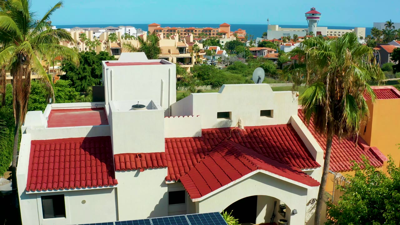 Elevated view over red roof tiles, white walls, palms, solar panels, distant buildings, and ocean.