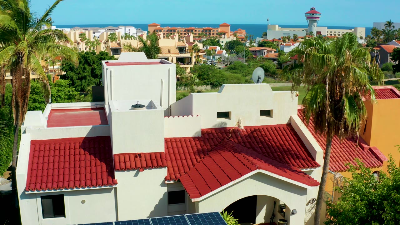 At 0:02, the San Jose Del Cabo rooftop view shows the red tiles and white walls centered below palms, with greenery, a distant tower, low buildings, ocean, blue sky, and no visible people.