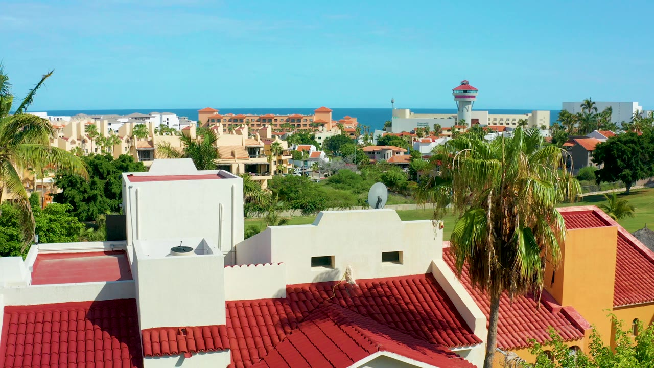 Elevated view in San Jose del Cabo of white residential buildings with red tile roofs, palms on both sides, distant homes, blue ocean horizon, and no visible people.