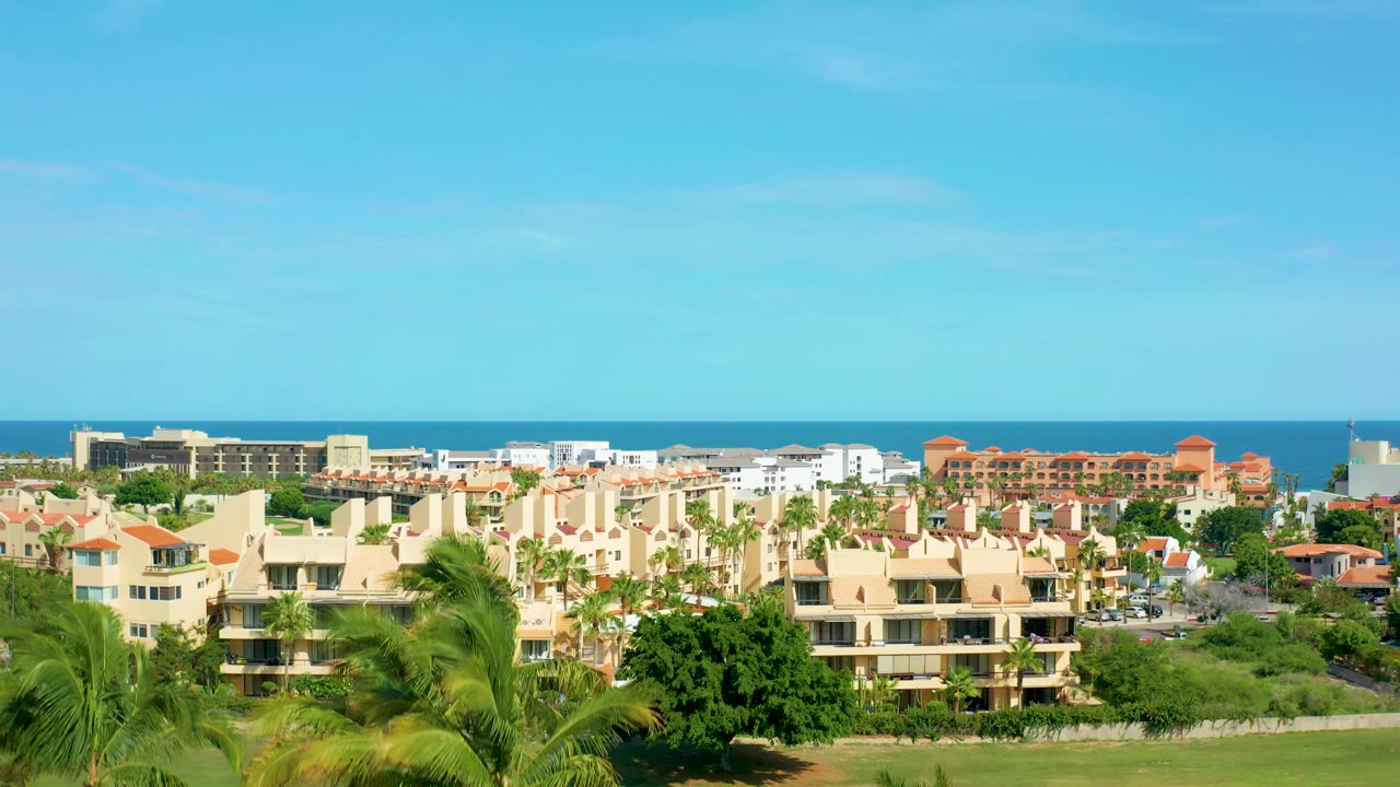 A wide San Jose Del Cabo view shows low-rise coastal housing, orange roof accents, green palms, and a thin blue ocean line under clear sky, with no visible people.