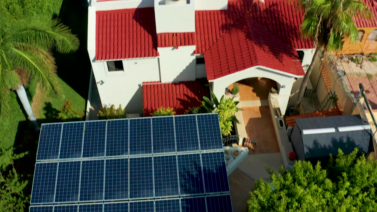 Overhead frame in San Jose Del Cabo showing blue solar panels beside a white house with a red tile roof, palm greenery, and no visible people.