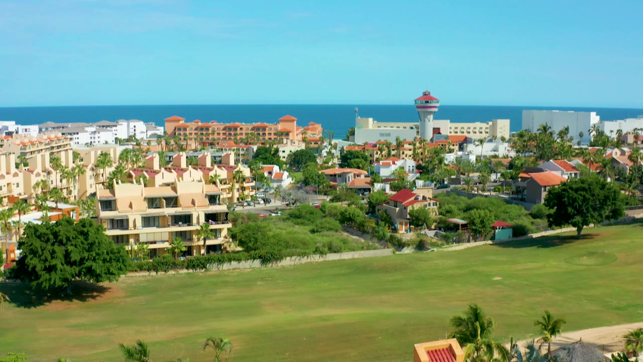 Wide aerial frame over San Jose Del Cabo with blue ocean and sky in the background, resort buildings beyond a large green lawn, red tile roofs, palms, and no visible people.