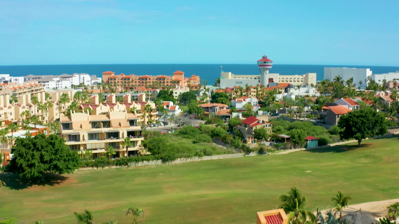 San Jose Del Cabo aerial view holds on the ocean horizon and resort district, with cream buildings, red roofs, palm trees, open grass, and no visible people.