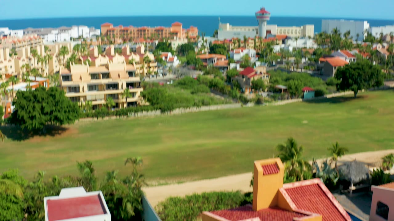 The aerial view in San Jose Del Cabo drops lower, shifting the green lawn and nearby red-roofed resort structures closer to the foreground, with no visible people.