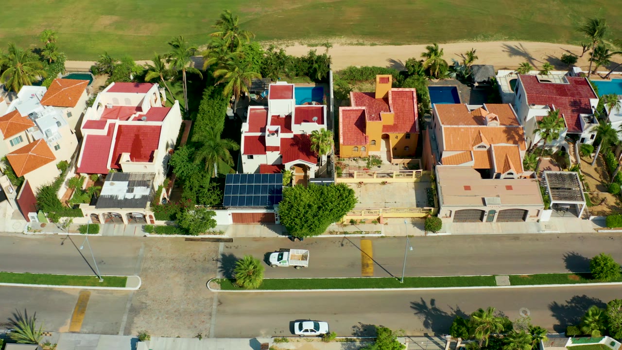Aerial frame over San Jose Del Cabo homes with the white car farther along the road, red roofs and a blue pool visible above the street, and no visible people.