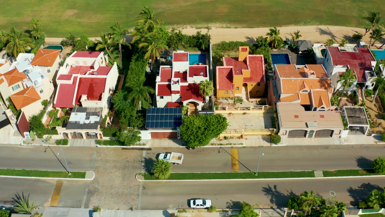 San Jose Del Cabo aerial view with the white vehicle near the lower road area, colorful rooflines, palm shadows, solar panels, and no visible people in the frame.