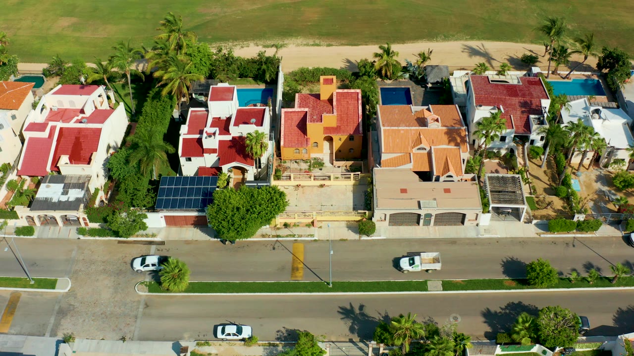 Aerial frame over San Jose del Cabo homes with red roofs, palm trees, blue pools, a paved street with white vehicles, beachside greenery at the top, and no visible people.