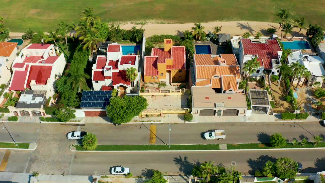 High aerial view in San Jose del Cabo showing white cars spaced along the street, colorful rooflines, palms, walled lots, pools, and no visible people.