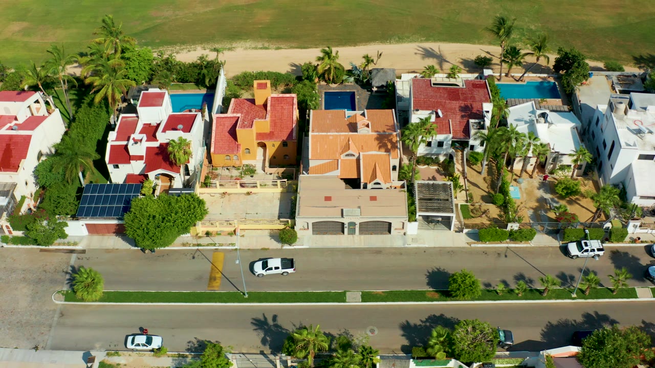 Overhead aerial frame in San Jose Del Cabo showing colorful beachfront homes, palm trees, pools, a sandy strip beyond the houses, a road in the foreground, and no visible people.