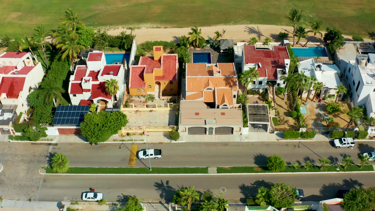 Aerial frame over San Jose Del Cabo homes with the beachside green area at the top, pools and palms between houses, white cars on the road below, and no visible people.