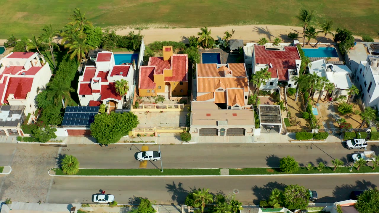 Overhead shot of coastal homes with red roofs, pools, palms, and a road.