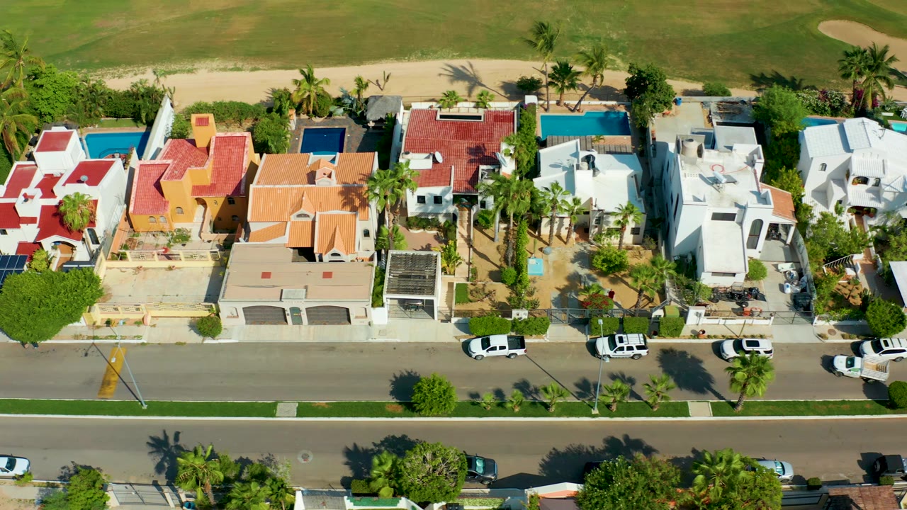 Aerial view of villas with pools, palms, solar panels, and parked cars along a road.