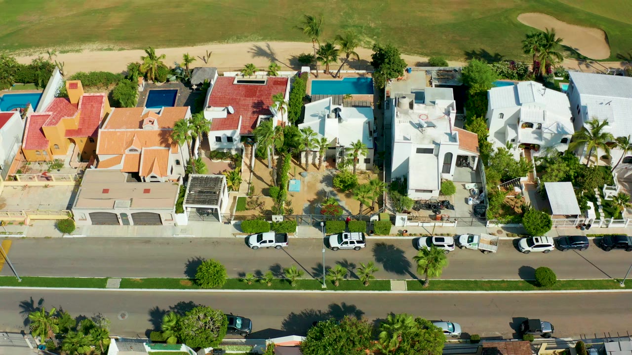 Overhead aerial frame in San Jose Del Cabo showing a row of residential buildings, parked cars, palm trees, pools, a green golf fairway, and a sand trap, with no visible people.