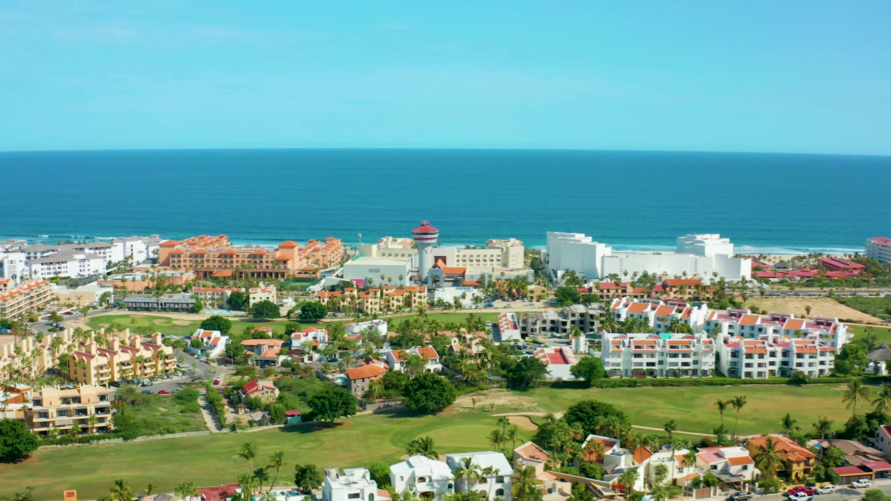 Aerial frame over San Jose Del Cabo showing red-roofed residences, white buildings, green foreground space, blue ocean, clear sky, and no visible people.