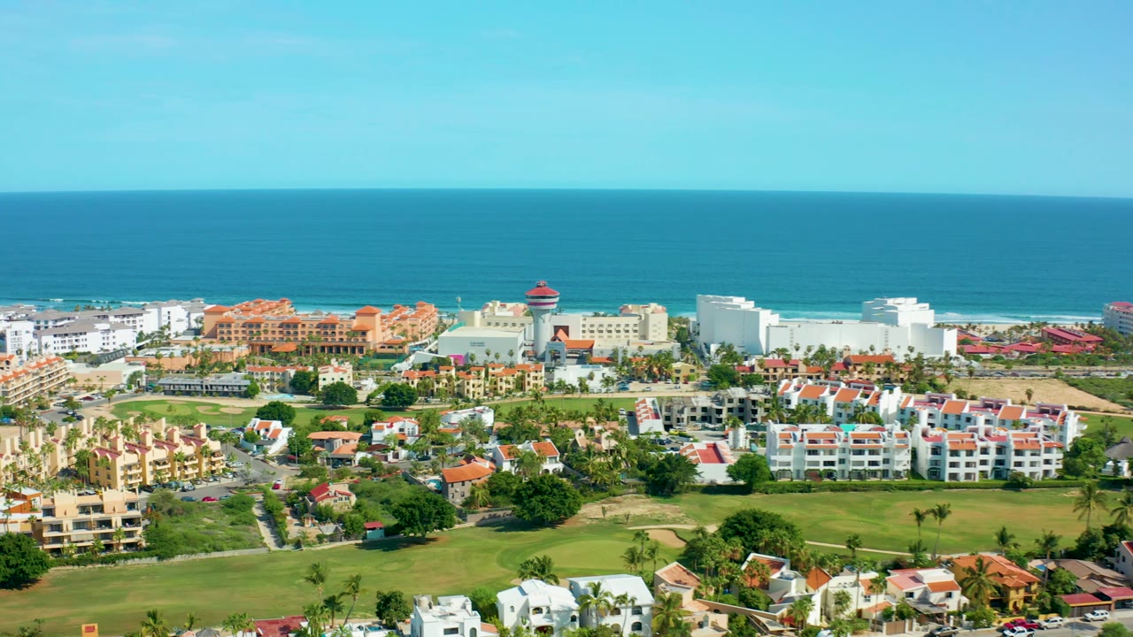 Aerial view in San Jose Del Cabo with white coastal buildings, red tile roofs, landscaped green areas, palm trees, ocean background, and no visible people.