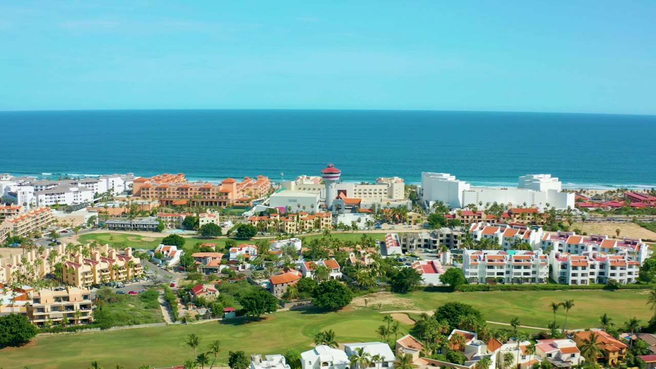 San Jose Del Cabo aerial frame with tan and white residences, palm trees, open green space, a straight ocean horizon, and no visible people.