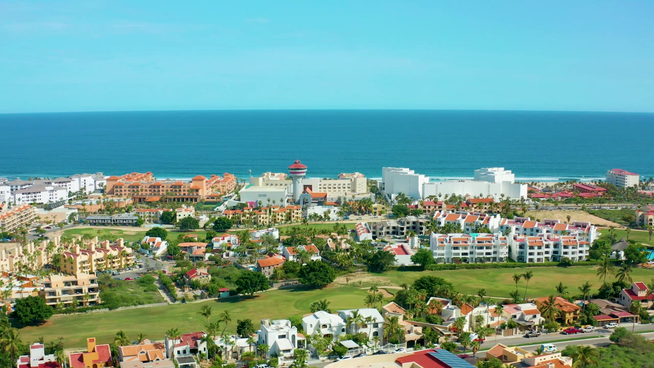 Wide aerial view over San Jose Del Cabo coastal homes and green lawns toward a blue ocean horizon, with no visible people.