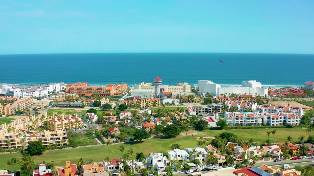 San Jose Del Cabo aerial frame with low buildings and green spaces below the ocean horizon as a small bird appears high in the clear sky, with no visible people.
