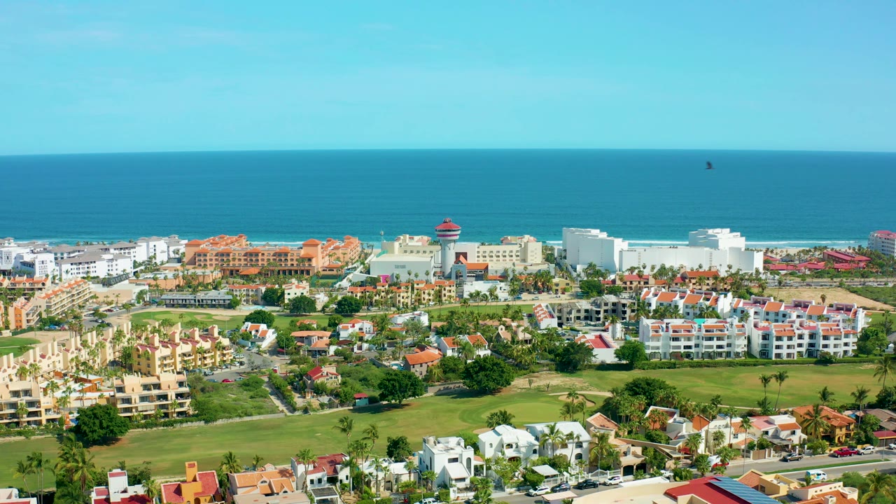 Wide San Jose Del Cabo aerial view with beachfront buildings, residential blocks, green lawns, blue ocean, clear sky, and no visible people.