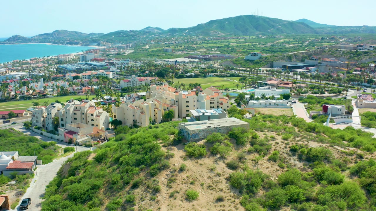 Wide aerial view of residences, roads, green areas, hills, and coastline.
