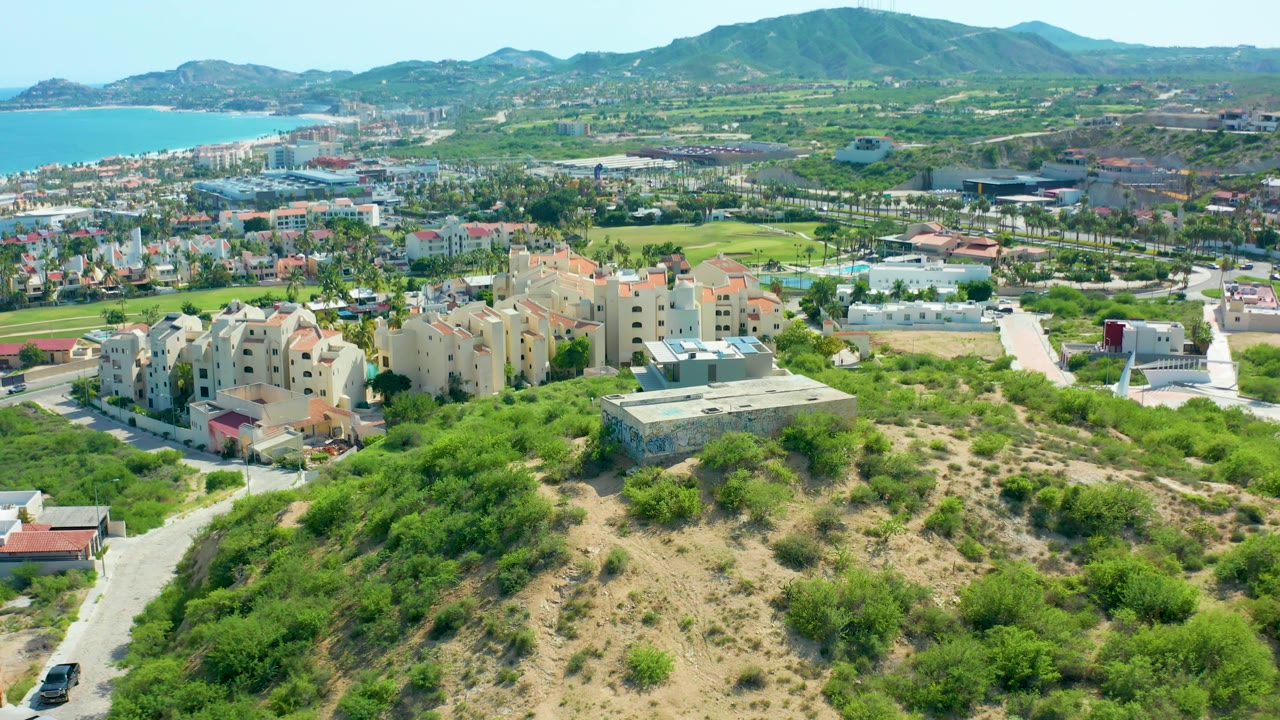 San Jose Del Cabo aerial frame with a larger foreground scrub hill, beige apartment-style buildings, pale roads, green corridors, ocean at upper left, mountains behind, and no visible people.