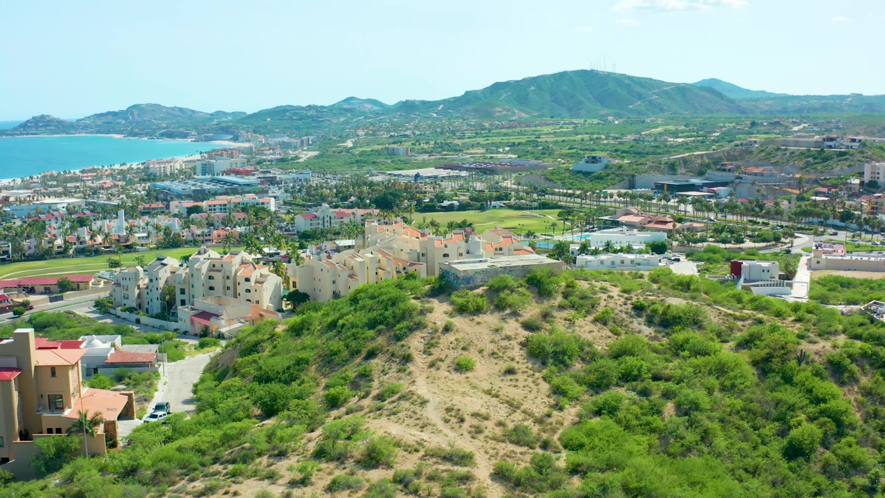 Second aerial frame in San Jose Del Cabo showing the same hillside foreground, clustered residential buildings, blue ocean on the left edge, distant mountains, and no visible people.