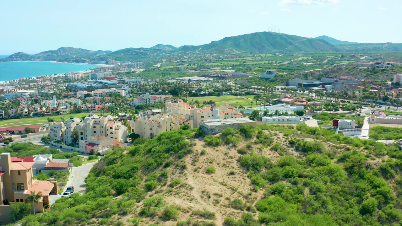 Third frame from the aerial view over San Jose Del Cabo, with hillside brush filling the lower frame, cream housing and roads in the middle distance, ocean to the left, and no visible people.