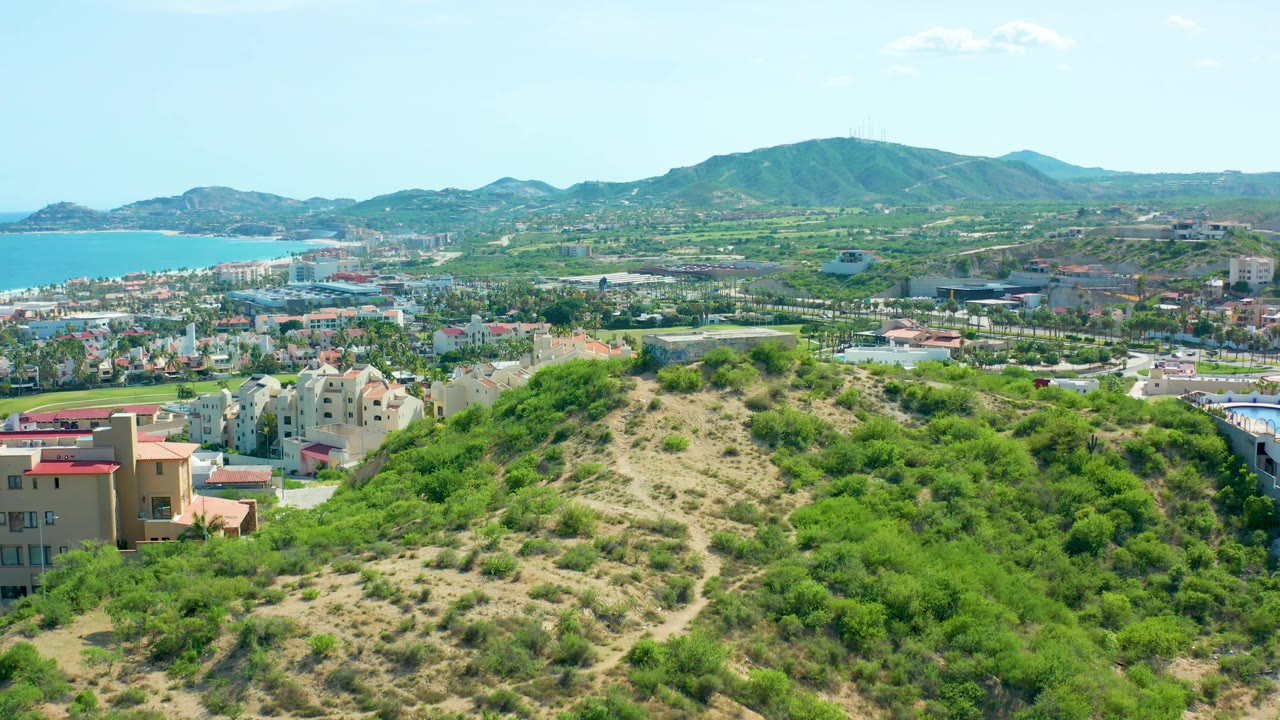San Jose del Cabo hillside view with the sandy ridge centered, dense green vegetation on both sides, coastal water to the left, urban blocks below, distant mountains, and no visible people.