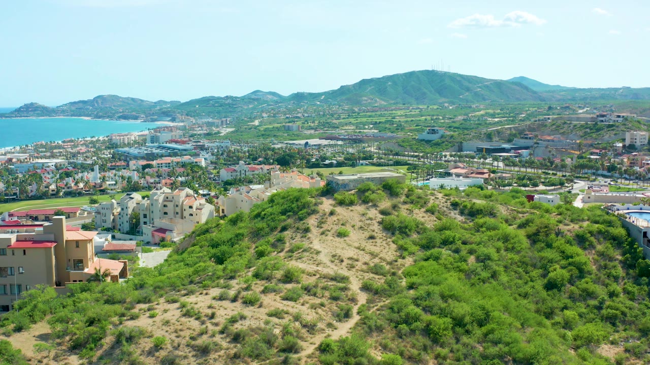 A bright San Jose del Cabo panorama shows brushy green hillside in the foreground, beige and white buildings in the midground, blue ocean on the left edge, mountains across the horizon, and no visible people.