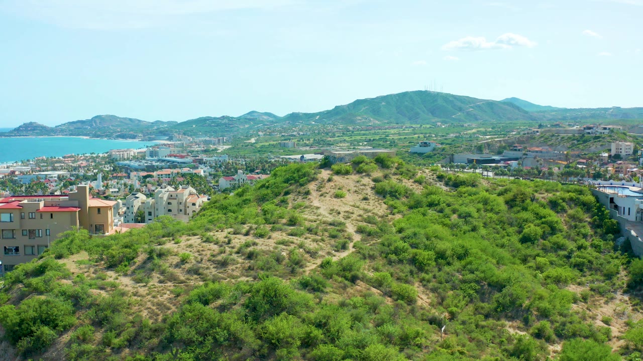 Opening aerial frame over San Jose Del Cabo, Mexico, with dense green hillside vegetation in front, low-rise city buildings below, blue sea at left, mountains on the horizon, and no visible people.