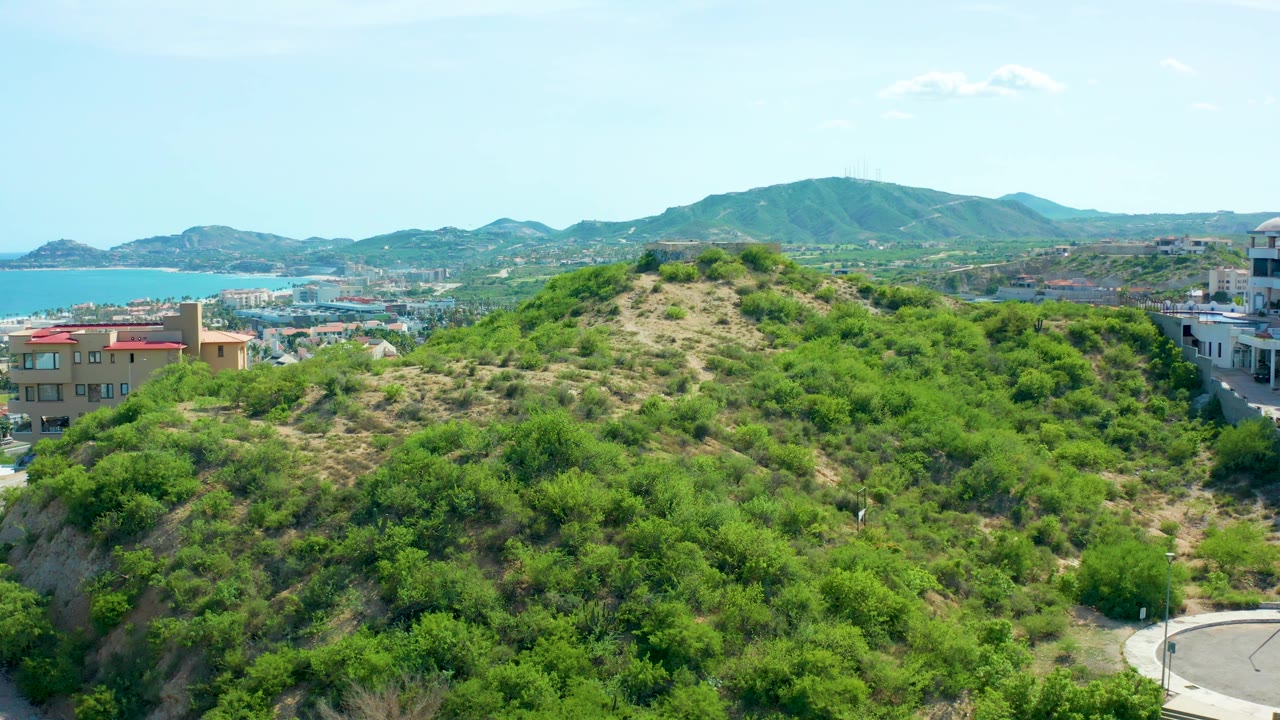 San Jose Del Cabo hillside ridge fills the foreground while tan buildings, the ocean, and rounded mountains remain visible beyond, with no visible people.