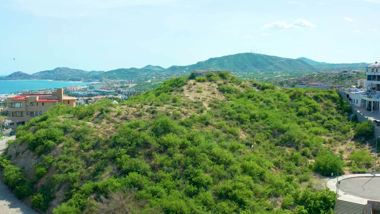San Jose Del Cabo hillside covered in green scrub with buildings at the edges, blue water and distant mountains beyond, and no visible people at the start of the clip.