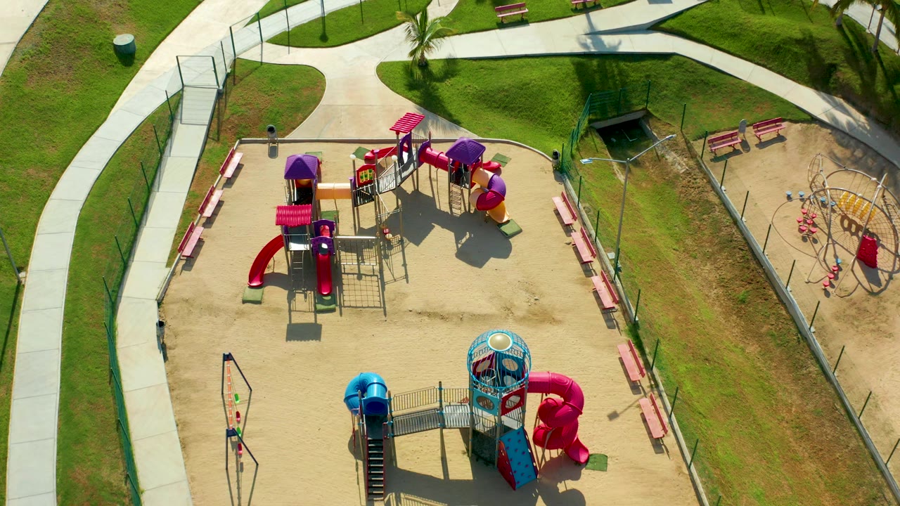 Overhead frame in San Jose Del Cabo showing a sandy playground with red and purple slides, blue equipment, benches, curved concrete paths, grass, and a small person on the left path.
