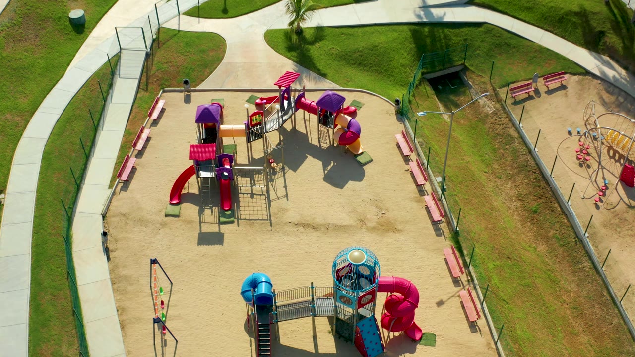 High overhead San Jose Del Cabo frame showing the play area shifted lower in the composition, with red and purple structures, a blue slide, benches, fencing, grass, and a small person on the left path.