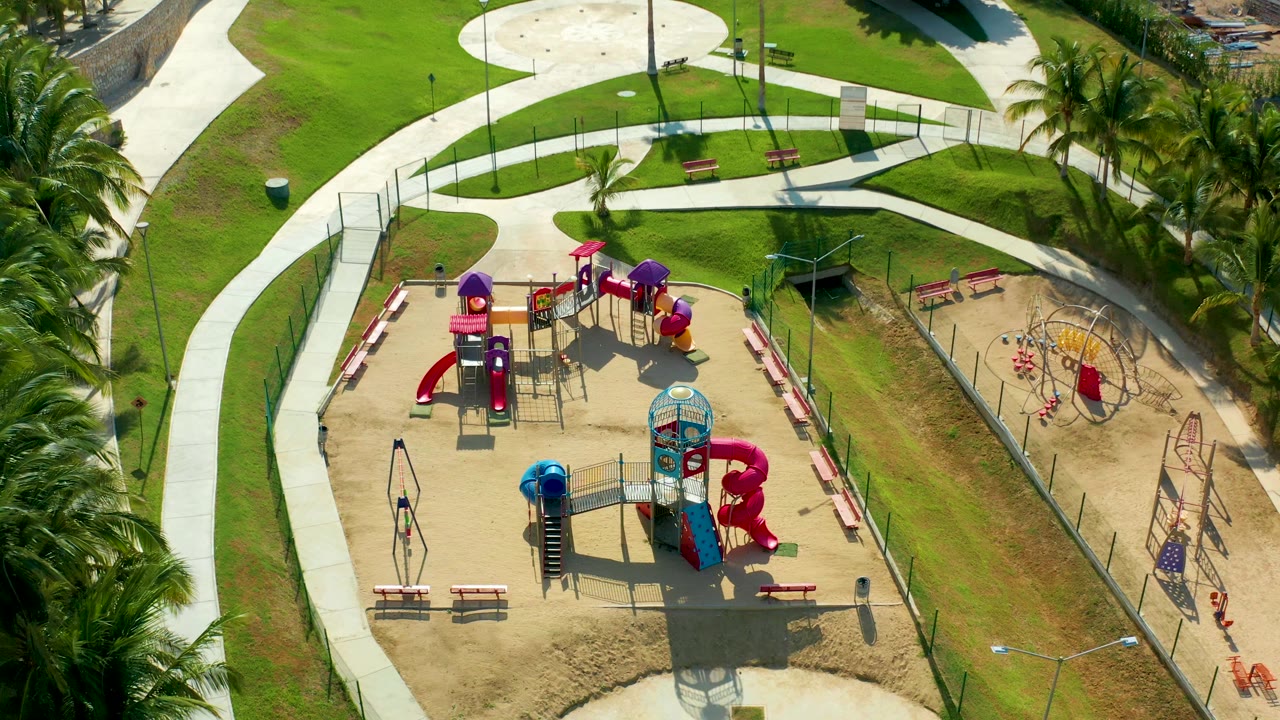 High drone view in San Jose Del Cabo showing the playground slightly closer, with red slides, purple roofs, blue tunnel elements, benches, lawn, palms, fencing, and no visible people.