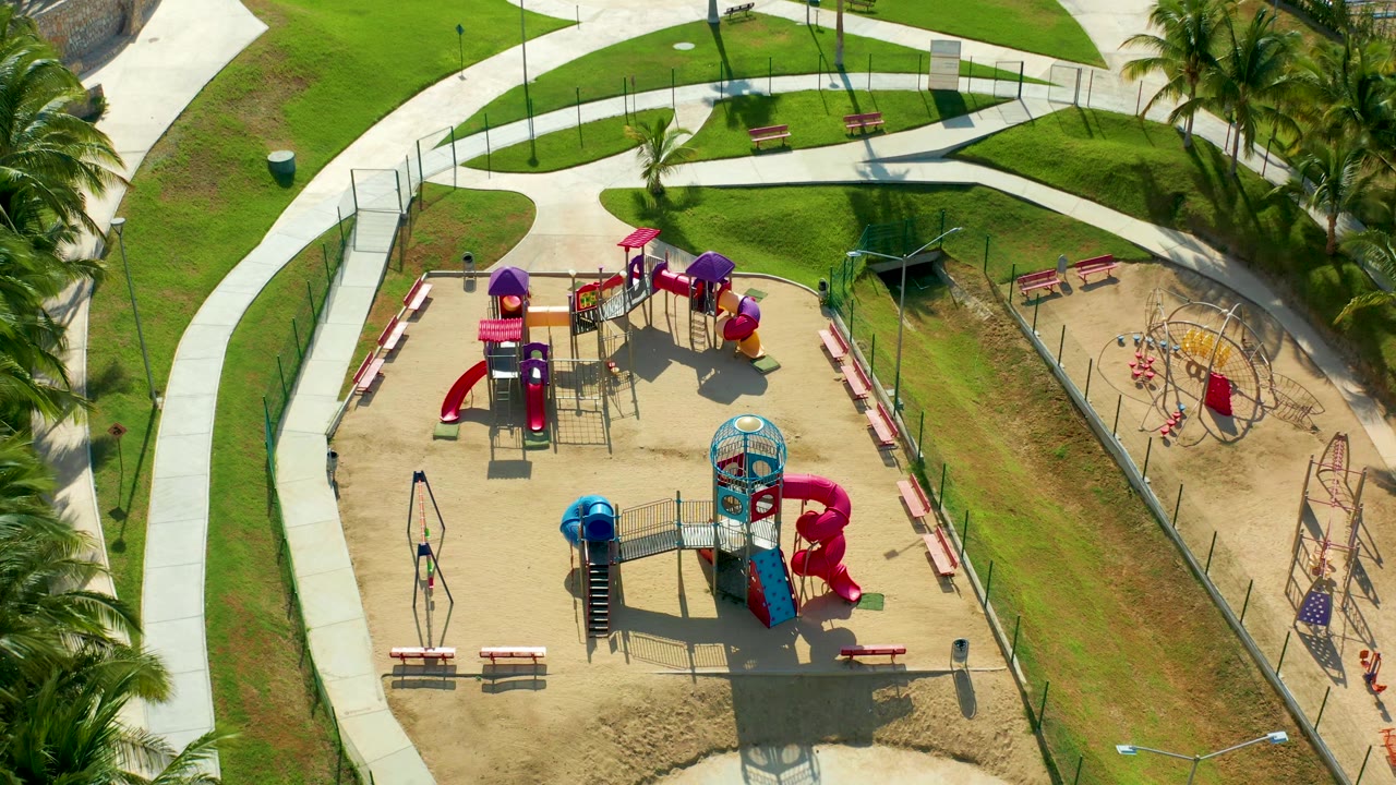 Overhead view of playground equipment, benches, grass, palms, and a fenced play area.