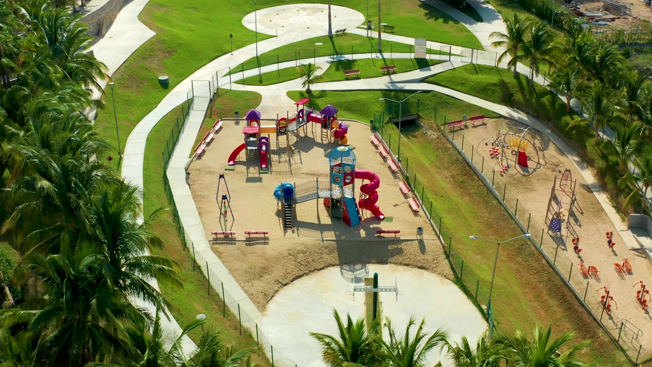 Aerial frame over a sandy playground in San Jose Del Cabo, Baja California Sur, Mexico, with red and blue slides, swings, benches, curved paths, green grass, palms, and small visible people near the right side.