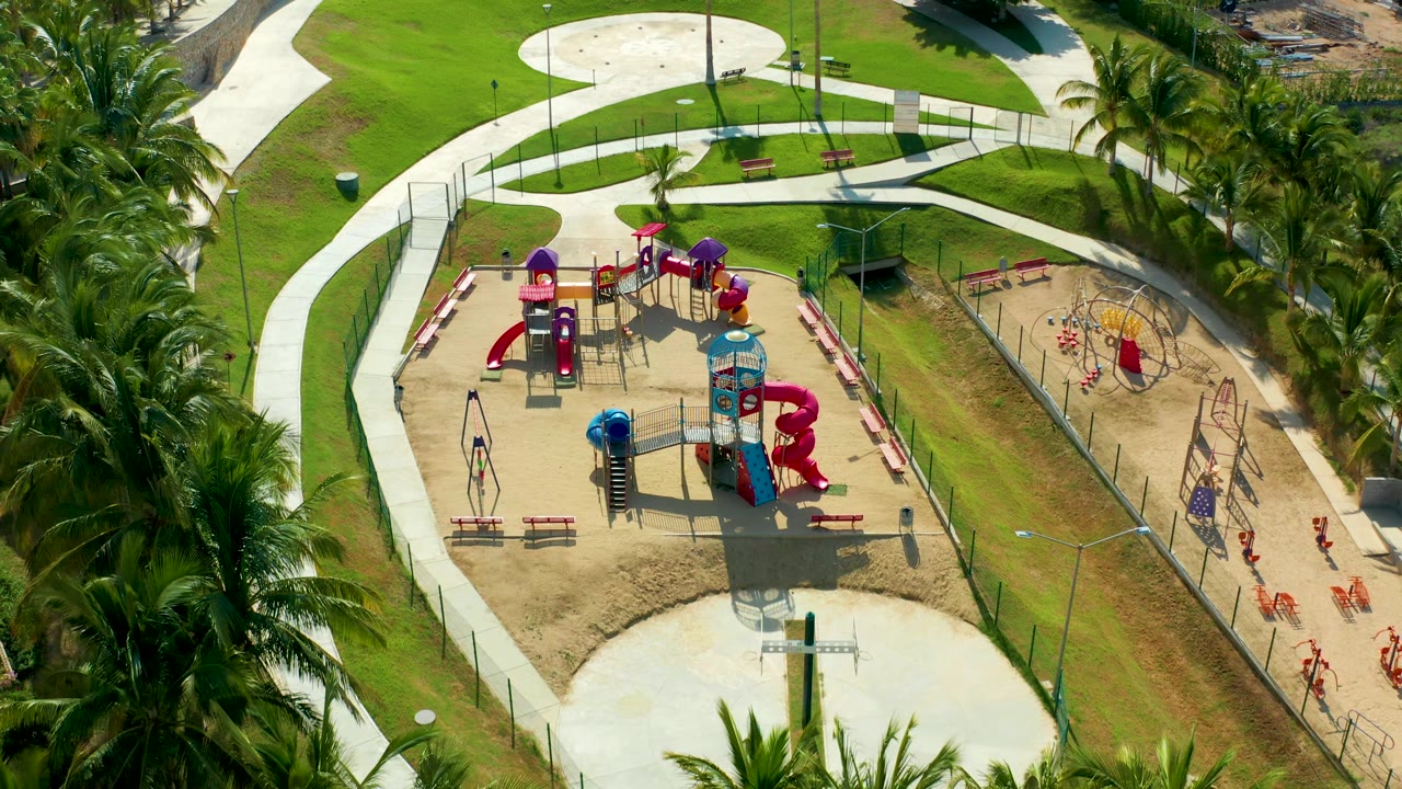 Overhead San Jose Del Cabo frame with red slides, a blue tower, swing sets, sandy ground, a green lawn, curved paths, palms on the right, and tiny people visible near the fenced play zone.