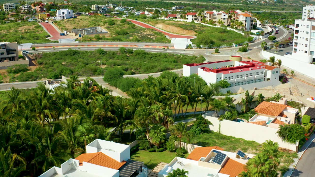 San Jose Del Cabo aerial view with palms and walled homes in the lower frame, a red-roofed white building near the road, open lots behind, and no visible people.
