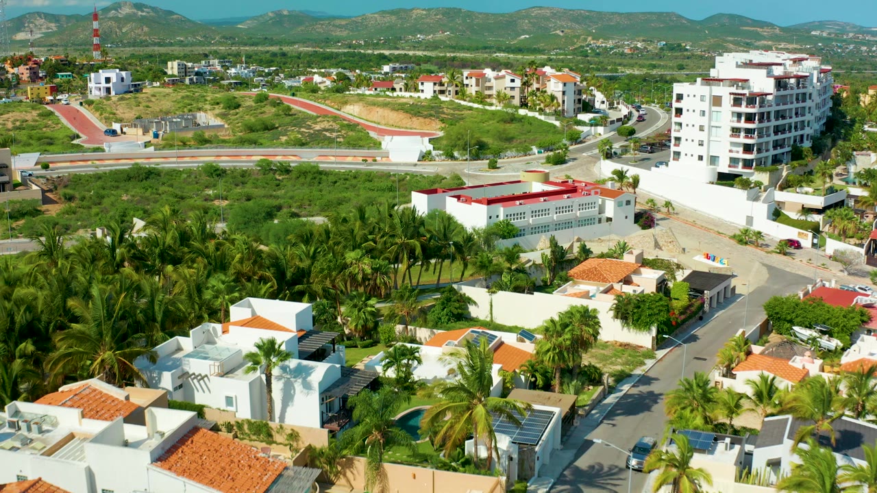 Opening aerial frame over San Jose del Cabo showing white residential buildings, orange tile roofs, palms, a curving road, distant hills, and no visible people.